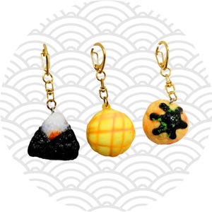 Könnte beinhalten: Drei Schlüsselanhänger mit Essensmotiven. Einer ist ein schwarz-weißes Onigiri, ein weiterer eine gelbe Cantaloupe-Melone und der dritte ein runder, orangefarbener Kuchen mit schwarzem Design. Goldfarbene Hardware.