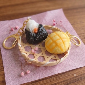 May include: Two food-themed keychains featuring a triangular onigiri with a dark seaweed wrap and a round, golden-brown melon pan. Both items are attached to gold-toned metal lobster clasps and are resting inside a small, round woven bamboo basket.
