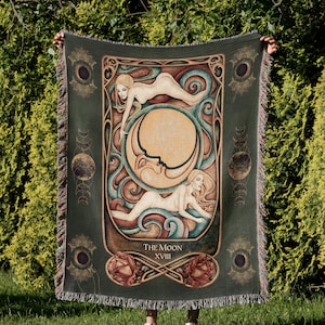 Peut inclure: Une couverture en tissu tissé avec un fond vert et une image colorée de la carte de tarot de la Lune. La carte représente deux figures nues et une lune croissante avec un visage. Le texte "THE MOON XVIII" est imprimé en bas de la carte.