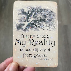 May include: A card featuring a quote from the Cheshire Cat from Alice in Wonderland. The quote reads "I'm not crazy. My Reality is just different from yours." The card has a glittery background and a black and white illustration of the Cheshire Cat.