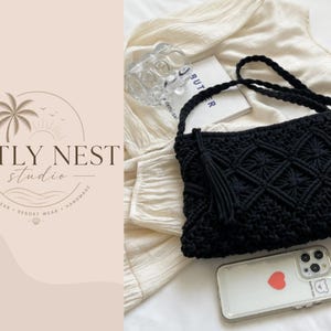 Puede incluir: Un bolso bandolera de macramé negro con un tejido de patrón de diamantes y una borla lateral. El bolso tiene una correa trenzada y está colocado junto a un objeto de vidrio transparente, un libro blanco y un smartphone con un adhesivo de corazón.