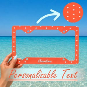 May include: A rectangular orange license plate frame featuring a white polka dot pattern and the name Carolina printed in white script on the bottom edge. A circular inset shows a close-up of the orange surface with white polka dots.