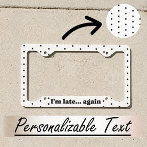 May include: A white rectangular license plate frame featuring a black polka dot pattern. The bottom center of the frame displays the black text "I'm late... again". A circular inset at the top right shows a magnified view of the black dot pattern.