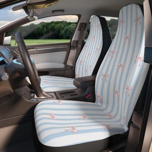 May include: Car seat covers with a light blue and white striped pattern, adorned with pink bows. The covers are visible inside a car, with the steering wheel and dashboard in view. The design adds a touch of whimsy to the car's interior.