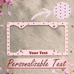 May include: A rectangular license plate frame featuring a light pink background with a pattern of small dark red polka dots. The bottom section of the frame includes the text Your Text in a dark red font, with four mounting holes at the corners.