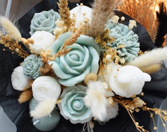 Könnte beinhalten: Ein Blumenstrauß aus salbeigrünen und weißen blütenförmigen Kerzen, arrangiert mit getrockneten Weizenhalmen, flauschigem Pampasgras und kleinen gelben Füllblumen. Das Arrangement ist in schwarzes Papier eingewickelt, das einen Kontrast zu den hellen Elementen bildet.