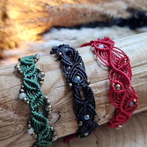Könnte beinhalten: Drei Makramee-Armbänder in Grün, Schwarz und Rot, jedes mit komplexen Knotenmustern, kleinen Metallperlen und hellen, kugelförmigen Akzenten, die in das geflochtene Kordeldesign eingearbeitet sind und nebeneinander auf Holz liegen.