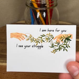 Könnte beinhalten: Ein rechteckiger weißer Aufkleber mit einer Illustration von zwei Händen, die aufeinander zu greifen und durch eine grüne Ranke verbunden sind. Der Text "I am here for you" steht über der Ranke, "I see your struggle" steht darunter.