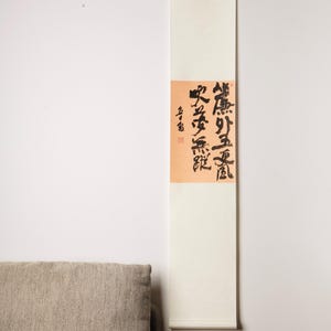 Könnte beinhalten: Eine vertikale Schriftrolle mit japanischer Kalligraphie in schwarzer Tinte auf cremefarbenem Hintergrund. Die Kalligraphie befindet sich auf einem pfirsichfarbenen rechteckigen Feld. Die Schriftrolle hängt an einer weißen Wand mit einem beigen Sofa in der linken unteren Ecke.