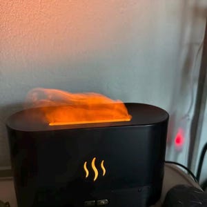 Peut inclure: Un diffuseur d'huiles essentielles noir de forme rectangulaire aux bords arrondis. L'appareil présente un effet lumineux orange imitant une flamme s'échappant de la fente supérieure et une icône de flamme éclairée sur le panneau avant.