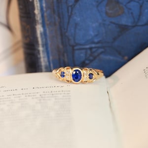 May include: A gold ring with a central oval blue gemstone and smaller blue stones on either side. The ring is set on an open book with text, with a blue book in the background.