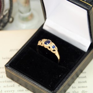 Könnte beinhalten: Ein goldener Ring mit drei ovalen blauen Edelsteinen und dekorativen Filigran-Details, präsentiert in einer schwarzen Schmuckschatulle mit weißem Innenraum. Der Ring liegt vor einem offenen Buch.