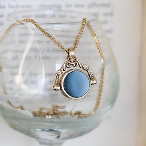 Può includere: Una collana con pendente dorato con una pietra centrale rotonda azzurra. Il pendente è sospeso a una delicata catena ed è esposto all'interno di un bicchiere trasparente. Lo sfondo include un libro aperto sfocato.