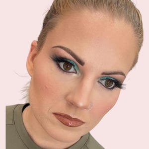 Puede incluir: Primer plano de un rostro con un maquillaje de ojos llamativo. Los ojos se acentúan con sombra de ojos turquesa, delineador oscuro y largas pestañas negras. Los labios son de un color brillante, de tono marrón. La piel tiene una tez natural y rosada.