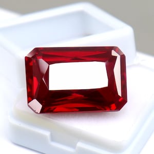 May include: A vibrant, deep red gemstone cut into a rectangular shape with faceted edges. The stone features a polished, reflective surface that catches the light, creating bright highlights across its top facets. It is displayed in a white box.