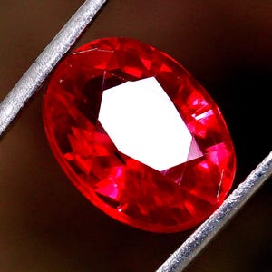 Könnte beinhalten: Ein leuchtend roter Edelstein im Ovalschliff, der von einer silberfarbenen Metallpinzette gehalten wird. Der Stein hat eine facettierte Oberfläche, die das Licht reflektiert und in der Mitte einen hellen weißen Glanzpunkt erzeugt, während die umliegenden Facetten einen tiefen, gesättigten Rotton aufweisen.