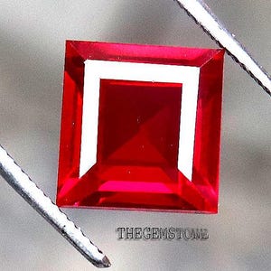 May include: A square-cut, vibrant red gemstone held by metal tweezers. The stone features a faceted surface with a distinct white square reflection in the center. The text THEGEMSTONE is printed in small black letters directly below the red gemstone.