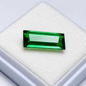 May include: A vibrant, emerald green gemstone cut into a long, rectangular baguette shape with sharp, faceted edges. The stone displays a clear, transparent quality and is positioned centrally on a white, textured square display pad.
