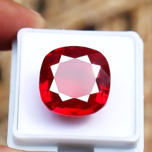 Op de afbeelding: Een levendige rode edelsteen met een kussensnede, gepresenteerd in een witte behuizing. De edelsteen heeft een centraal sterrenpatroon met witte facetten, wat een opvallend contrast creëert met de dieprode tint. De edelsteen zit in een witte plastic behuizing.