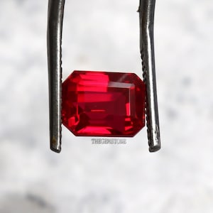 May include: A bright red, rectangular gemstone with multiple facets is held by metal tweezers. The gemstone has a reflective surface. The text "THEGEMSTONE" is visible on the gemstone.