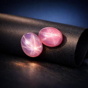 Op de afbeelding: Twee ovale, roze ster saffieren. Elke edelsteen toont een zesstralig sterpatroon over het oppervlak. De edelstenen zijn geplaatst tegen een donkere, getextureerde achtergrond, met een blauwe gradiënt achtergrond.