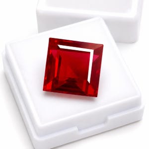 Puede incluir: Una piedra preciosa de color rojo vibrante con corte cuadrado y superficies facetadas que reflejan la luz, colocada sobre un inserto de espuma blanca dentro de una pequeña caja de almacenamiento de plástico blanco abierta con bordes definidos.