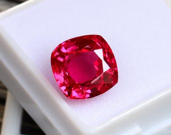 Puede incluir: Una gema roja vibrante de corte cojín con una superficie facetada que refleja la luz, exhibida dentro de una pequeña caja de almacenamiento cuadrada de plástico blanco con un inserto de espuma blanca. La gema muestra un color carmesí saturado.