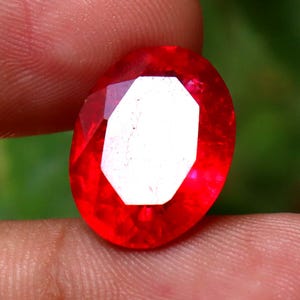 May include: A faceted, oval-shaped, bright red gemstone with a large, white, geometric center. The gem is held between fingers, with a blurred green background.