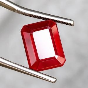 Könnte beinhalten: Ein leuchtend roter Edelstein im Rechteckschliff, der von einer silberfarbenen Metallpinzette gehalten wird. Der Stein hat eine facettierte Oberfläche mit scharfen Kanten und ein klares, transparentes Aussehen, das Licht auf seinen polierten, tiefroten Facetten reflektiert.