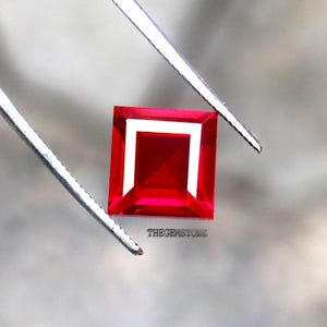 May include: A square-cut, vivid red gemstone with a white border, held by silver tweezers. The gemstone has a bright, polished surface. The text "THEGEMSTONE" is visible on the bottom right. This image is ideal for jewelry and gemstone listings.