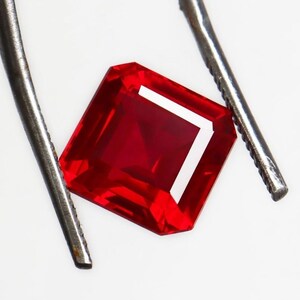May include: A square-cut, ruby-red gemstone is held by metal tweezers against a white backdrop. The gemstone's facets reflect light, highlighting its clarity and rich colour. The image showcases the gemstone's brilliance and the precision of its cut.