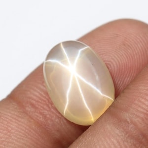May include: A polished, oval-shaped gemstone with a star effect. The gem is a creamy beige color, with a six-rayed star pattern radiating from the center. The star effect is created by light reflecting off of inclusions within the stone.