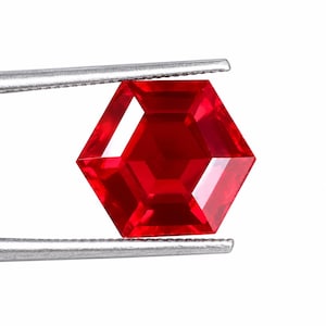 May include: A vibrant, transparent red gemstone cut into a precise hexagonal shape. The gemstone features multiple facets that reflect light, creating a brilliant appearance. It is held securely by a pair of silver-toned metal tweezers.