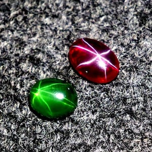 May include: Two gemstones: a vibrant green cabochon and a deep red cabochon. The green stone has a star effect, with lines radiating from a central point. The red stone displays a six-rayed star pattern. Both are oval-shaped and set against a textured background.