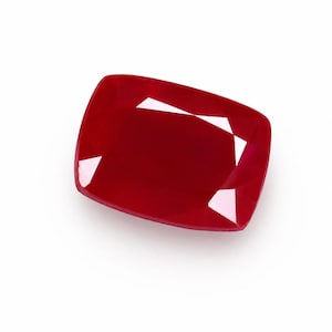 May include: A cushion-cut ruby gemstone with a deep, saturated red color. The stone features a faceted surface that reflects light, creating distinct geometric patterns across its top. The edges are rounded, and the overall shape is rectangular.