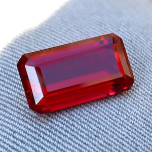 May include: A faceted, rectangular-cut, deep red gemstone. The gem has a polished surface with sharp edges and a transparent appearance. The color is a rich, vibrant red, with a slight internal depth. The gemstone is set against a textured, light gray fabric.