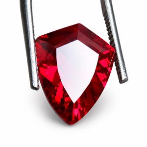 Puede incluir: Una gema facetada en forma de escudo de color rojo intenso. La piedra presenta una disposición compleja de facetas triangulares y trapezoidales que crean reflejos de luz nítidos y un centro claro y brillante. Está sujeta por unas pinzas de metal.