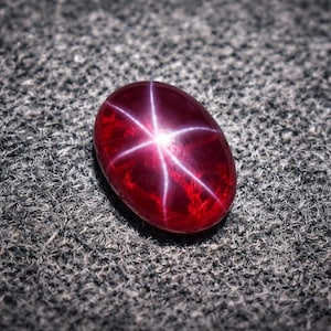May include: A polished, oval-shaped, deep red gemstone with a six-rayed star pattern. The star appears as bright white lines radiating from the center of the stone. The gem is set against a textured gray background.