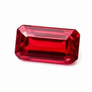 May include: A vibrant red gemstone cut into a rectangular emerald shape with faceted edges. The stone displays a deep, saturated crimson hue with internal light reflections visible across its polished, transparent surface against a white background.