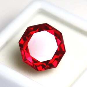 Könnte beinhalten: Ein leuchtend roter Edelstein im Achteckschliff mit zahlreichen Facetten, die das Licht reflektieren. Der Stein besticht durch einen brillanten karmesinroten Farbton und eine polierte Oberfläche, die ein geometrisches Design mit scharfen Kanten zeigt.
