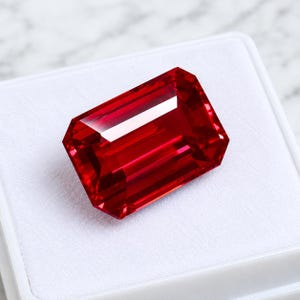 May include: A vibrant, deep red gemstone with a rectangular emerald cut, featuring sharp facets that reflect light. The stone is displayed resting on a white, square-shaped presentation box with a soft, textured interior lining.