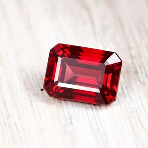 May include: A vibrant, deep red gemstone cut into a rectangular emerald shape. The stone features precise, multi-faceted edges that create sharp geometric reflections and a clear, transparent appearance with intense crimson color saturation throughout.