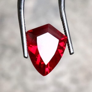 Könnte beinhalten: Ein facettierter, schildförmiger, tiefroter Edelstein, der von einer Metallpinzette gehalten wird. Der Edelstein hat eine helle, weiße, dreieckige Facette in der Mitte, die das Licht reflektiert. Die Gesamtform ist ein modifiziertes Dreieck mit mehreren Facetten.