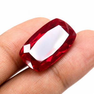 May include: A vibrant, deep red gemstone with a rectangular cushion cut and faceted surface. The stone displays high clarity and intense saturation, reflecting light across its polished edges while being held against a plain white background.