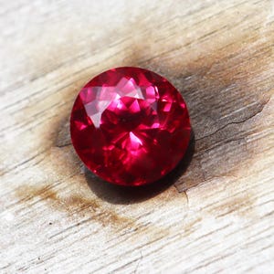 Pode incluir: Uma gema facetada, redonda, vermelho rubi. A gema é altamente reflexiva, com múltiplas facetas que capturam a luz. Está sobre uma superfície de madeira clara e envelhecida, realçando sua cor vibrante.