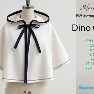 Könnte beinhalten: Ein weißer Kapuzenumhang mit dunkelblauem Saum und einer Bindung am Hals. Der Text rechts lautet: AtelierLiana PDF Sewing Pattern Dino Cape, Kid's sizes 2-3Y, 4-5Y, 6-7Y, 8-9Y, 10-11Y, 12-13Y, und Beginner friendly.