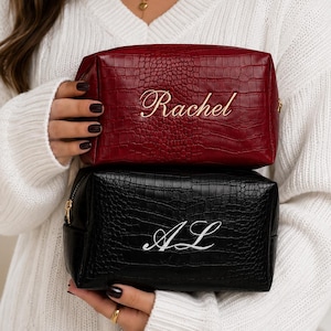 Puede incluir: Dos neceseres rectangulares con textura de piel de cocodrilo. El neceser superior es de color rojo oscuro con el nombre Rachel bordado en hilo dorado. El inferior es negro con las iniciales AL bordadas en hilo blanco. Ambos tienen cremalleras doradas.