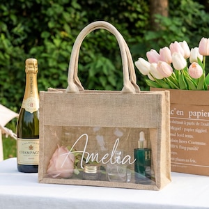 May include: A rectangular jute tote bag with a clear plastic front panel featuring the white cursive name Amelia printed on the center. The bag has two beige fabric handles and is positioned on a white tablecloth next to a bottle and a paper bag.