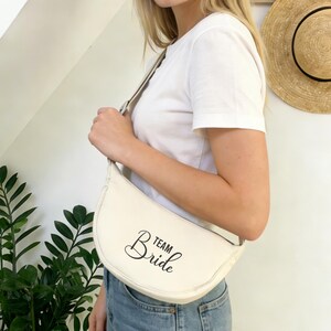 May include: Cream-colored, crescent-shaped bag with the text "Team Bride" in black script. The bag is worn over the shoulder. The person is wearing a white t-shirt and blue jeans. A straw hat hangs on the wall in the background.