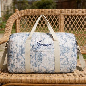 May include: A quilted duffel bag with a blue and white floral pattern, featuring cream-colored handles and a shoulder strap. The name "Joanna" is embroidered in blue on the front. The bag is resting on a wicker chair.